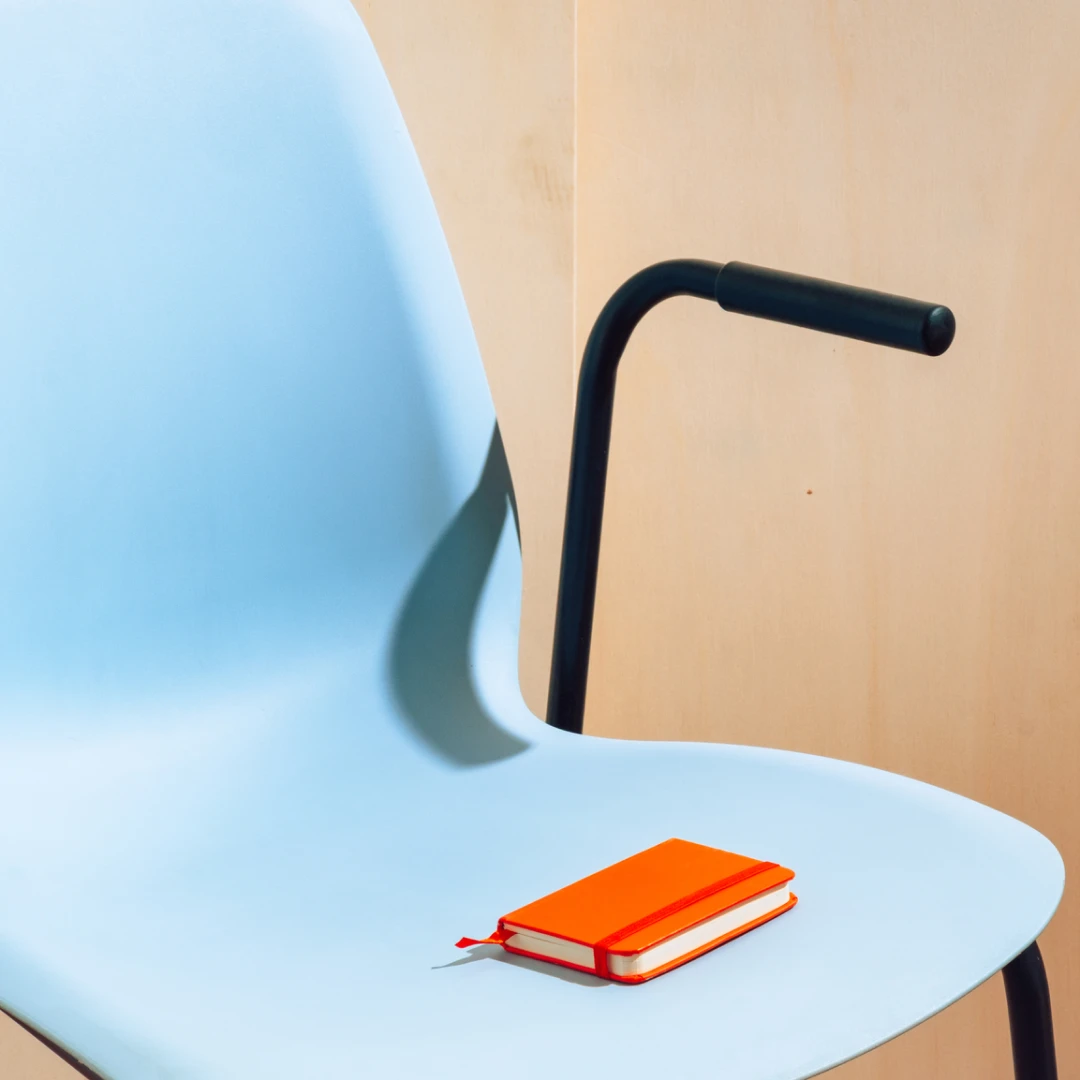 Imagen de un pequeño cuaderno de color naranja colocado sobre una silla de color azul celeste.