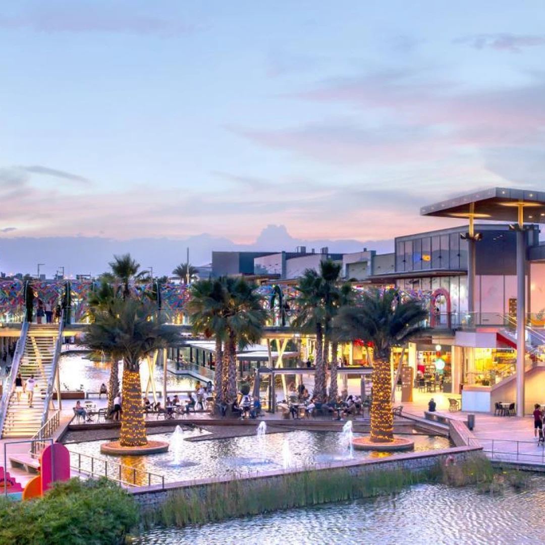 Complejo comercial junto al agua al atardecer, con palmeras iluminadas y fuentes